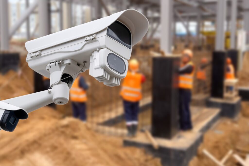Überwachungskamera an einem Mast vor einer Baustelle mit Bauarbeitern in Warnwesten