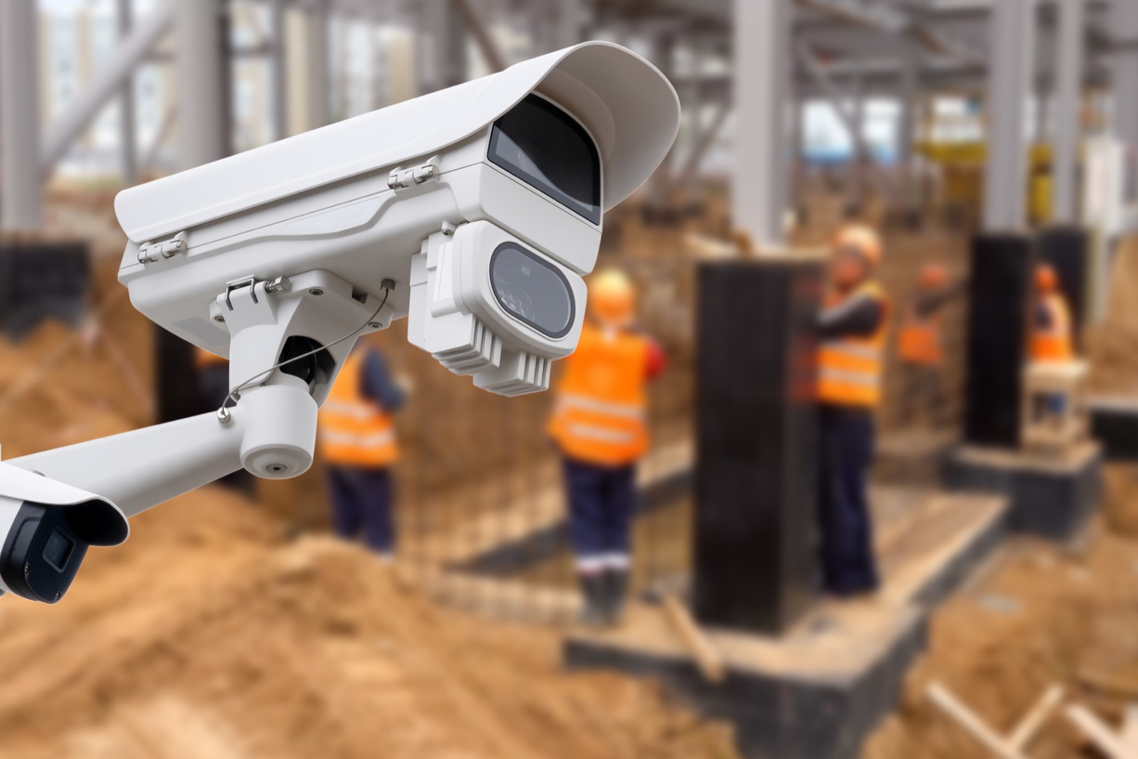 Überwachungskamera an einem Mast vor einer Baustelle mit Bauarbeitern in Warnwesten